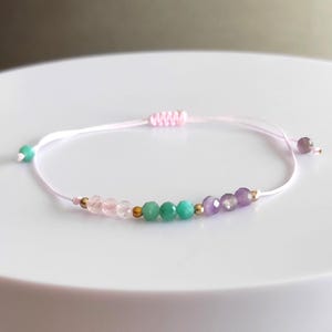 Könnte beinhalten: Ein zartes, verstellbares Armband in Rosa mit einer Reihe farbiger Edelsteinperlen. Die Perlen umfassen Rosatöne, Grün und Lila, durchsetzt mit kleinen goldfarbenen Perlen. Das Armband ist auf einem hellrosa Band aufgezogen.