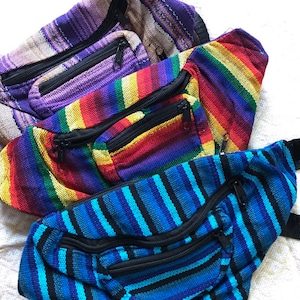 May include: Three colorful woven fanny packs with zippered pockets. The top pack is purple and white, the middle pack is rainbow striped, and the bottom pack is blue and black.