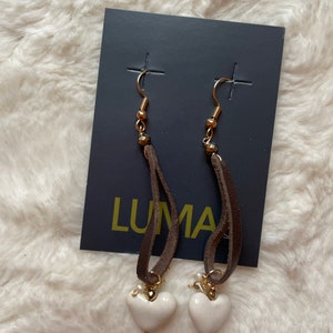 May include: A pair of white heart shaped earrings with brown leather cord and gold accents. The earrings are displayed on a black card with the word "LUMA" printed on it.