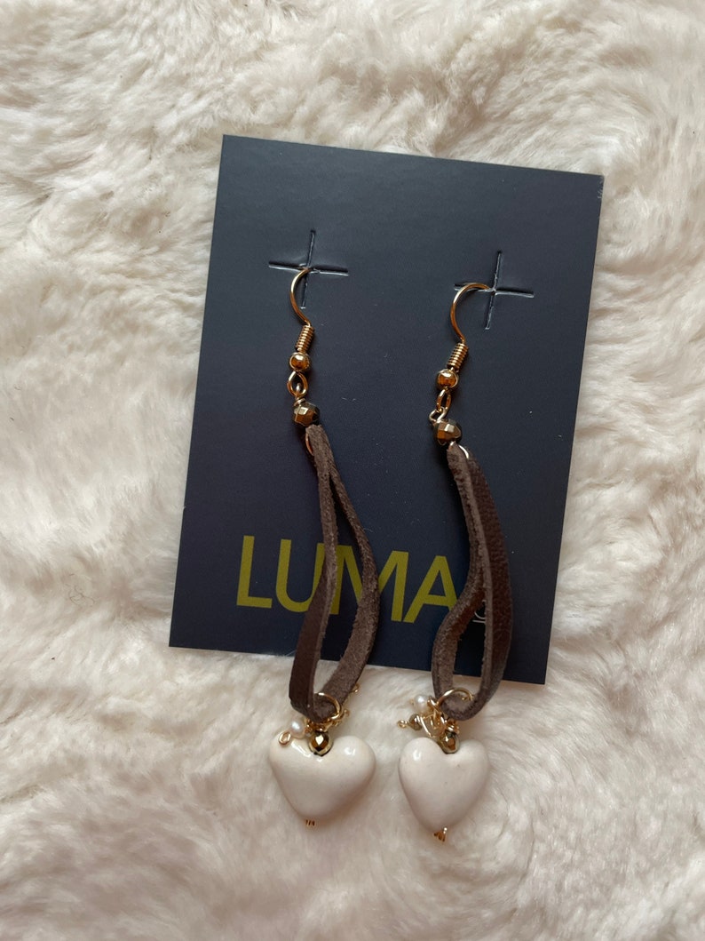 May include: A pair of white heart shaped earrings with brown leather cord and gold accents. The earrings are displayed on a black card with the word "LUMA" printed on it.