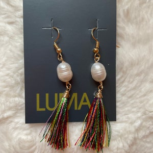 May include: A pair of gold earrings with a white pearl and a colorful tassel. The earrings are displayed on a gray card with the word "LUNA" printed on it.