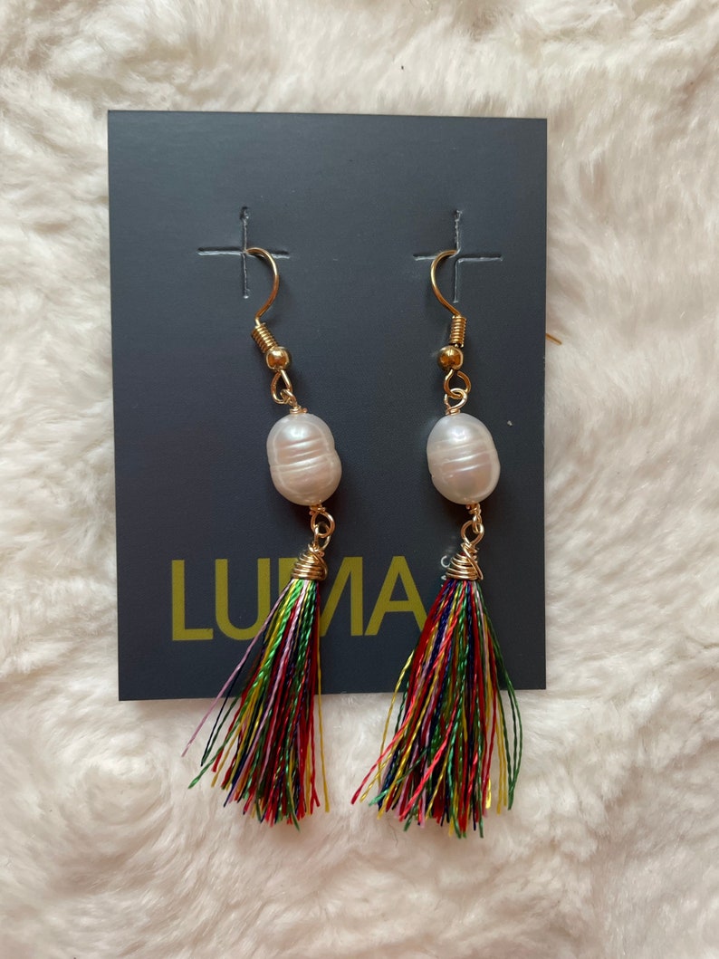 May include: A pair of gold earrings with a white pearl and a colorful tassel. The earrings are displayed on a gray card with the word "LUNA" printed on it.