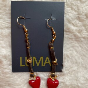 May include: A pair of dangle earrings with red heart charms. The earrings feature brown leather cords with gold beads and a gold wire wrapped around the cords. The earrings are displayed on a black card with the word "LUMA" printed on it.