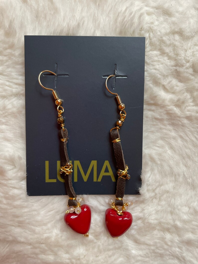 May include: A pair of dangle earrings with red heart charms. The earrings feature brown leather cords with gold beads and a gold wire wrapped around the cords. The earrings are displayed on a black card with the word "LUMA" printed on it.