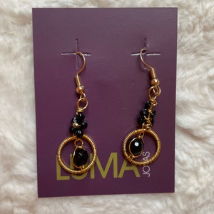 May include: A pair of gold hoop earrings with black beads and a larger black bead at the bottom. The earrings are displayed on a purple card with the word "LUMA" and "JOAS" printed on it.