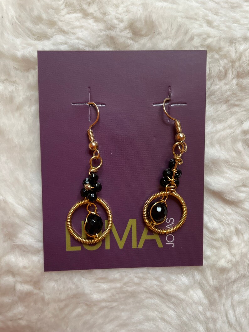May include: A pair of gold hoop earrings with black beads and a larger black bead at the bottom. The earrings are displayed on a purple card with the word "LUMA" and "JOAS" printed on it.