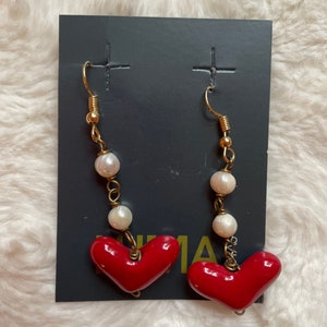May include: A pair of red heart dangle earrings with white pearl accents. The earrings are on a black card with the word "UMA" printed on it.