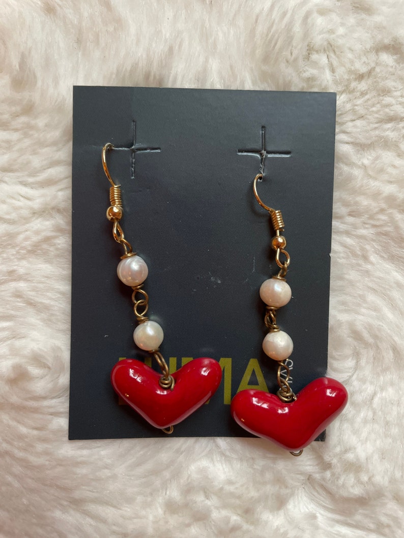 May include: A pair of red heart dangle earrings with white pearl accents. The earrings are on a black card with the word "UMA" printed on it.