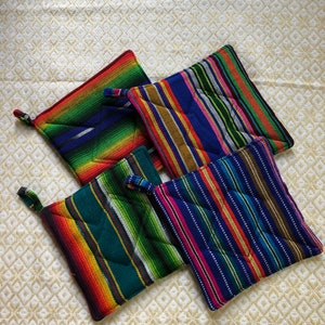 May include: Four colorful, square pot holders with striped patterns in various colors, including red, green, blue, and yellow. Each pot holder has a small loop for hanging. The pot holders are arranged on a cream-colored, patterned surface.