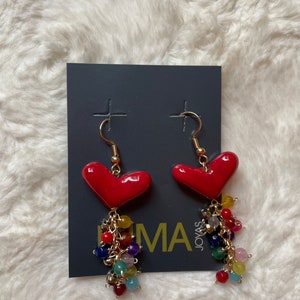 May include: A pair of red heart-shaped earrings with a dangling chain of colorful beads. The earrings are on a gray card with the text "Joyas Lima" printed on it.