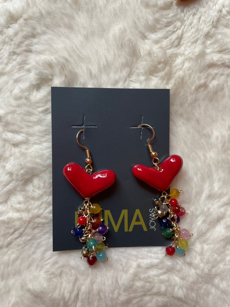 May include: A pair of red heart-shaped earrings with a dangling chain of colorful beads. The earrings are on a gray card with the text "Joyas Lima" printed on it.