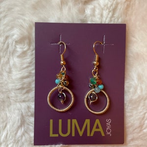 May include: A pair of gold hoop earrings with colorful gemstone beads. The earrings are on a purple card with the text "LUMA JOYAS".