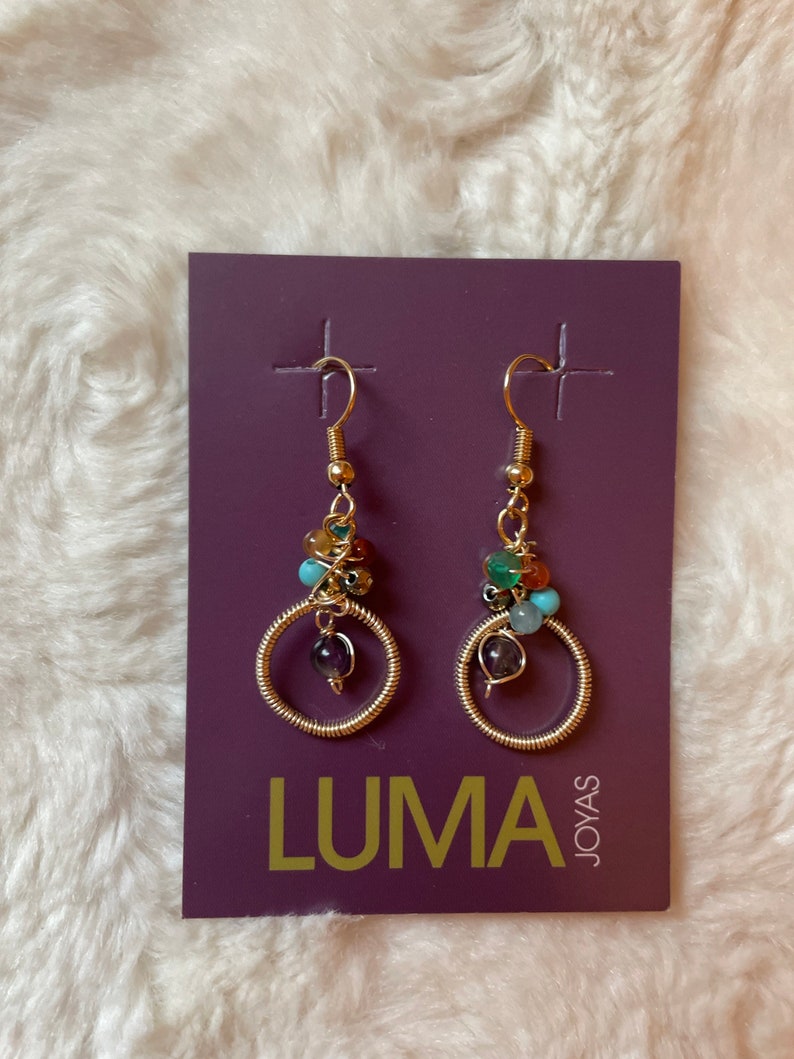 May include: A pair of gold hoop earrings with colorful gemstone beads. The earrings are on a purple card with the text "LUMA JOYAS".