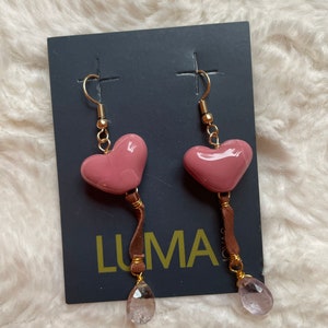 May include: A pair of pink heart-shaped earrings with brown leather tassels and clear crystal drops. The earrings are on a black card with the word "LUMA" printed on it.