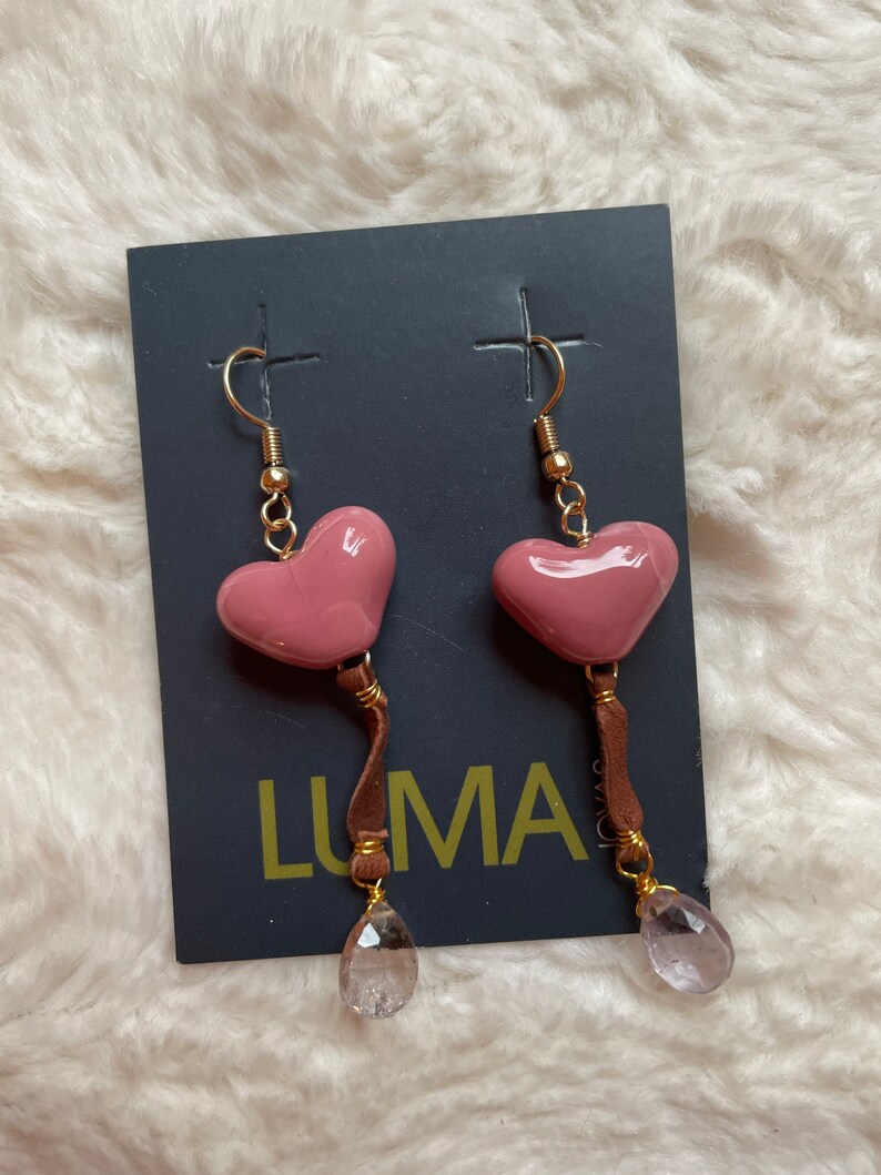 May include: A pair of pink heart-shaped earrings with brown leather tassels and clear crystal drops. The earrings are on a black card with the word "LUMA" printed on it.