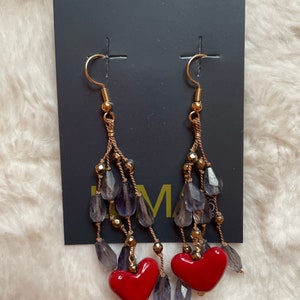 May include: A pair of dangle earrings with red heart charms. The earrings feature a gold chain with purple glass beads.