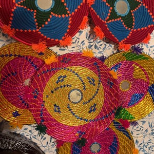 Op de afbeelding: Een collectie van zes ronde gevlochten wandkleden met centrale spiegels. De twee bovenste stukken tonen blauwe en rode sterpatronen met oranje kwastjes. De vier onderste stukken zijn voorzien van metallic gouden, roze en blauwe spiralen met kleine kwastjes.