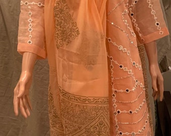 May include: Peach colored salwar kameez with a sheer dupatta. The kameez has a V-neckline and is decorated with small mirrors. The dupatta is also decorated with mirrors and has a floral print. The salwar is plain peach.
