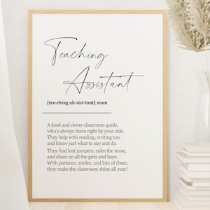 May include: A framed print with the words "Teaching Assistant" in elegant script. The definition of a teaching assistant is below the title. The print is in a light wood frame, with a vase of dried flowers and a stack of books in the background.
