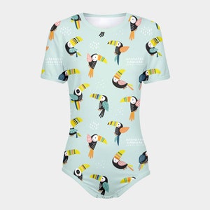 May include: A light blue baby onesie with a colorful toucan pattern. The toucans are depicted in a variety of poses, with some facing forward and others facing sideways. The onesie has short sleeves and a snap closure at the bottom.