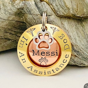 May include: Gold and copper dog tag with the text "I'm An Assistance Dog" and the name "Messi" engraved on it. A paw print is in the centre of the tag.