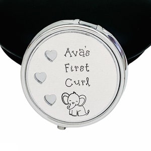 May include: A silver compact mirror with the text "Ava's First Curl" and a small elephant illustration. The mirror has three heart designs and a black, oval-shaped case.