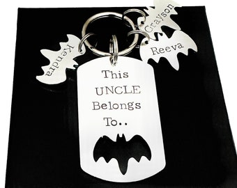 May include: A silver-toned metal keychain featuring a rectangular dog tag engraved with the text This UNCLE Belongs To.. and a bat silhouette. Three smaller bat-shaped charms are attached to the ring, each engraved with the names Kendra, Grayson, and Reeva.