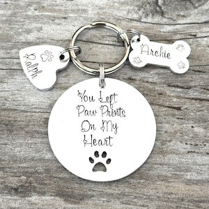 May include: Silver keyring with a heart charm engraved with "Ralph", a bone charm engraved with "Archie", and a round charm with the text "You left paw prints on my heart" and a paw print cutout.
