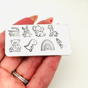 May include: A rectangular metal plate featuring black line drawings of a train, bunny, elephant, giraffe, teddy bear, dinosaur, rainbow, and rocket. The illustrations are simple and cartoon-like, suitable for children's designs.