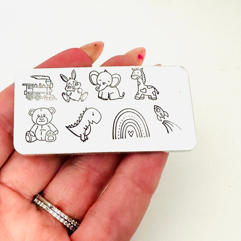 May include: A rectangular metal plate featuring black line drawings of a train, bunny, elephant, giraffe, teddy bear, dinosaur, rainbow, and rocket. The illustrations are simple and cartoon-like, suitable for children's designs.