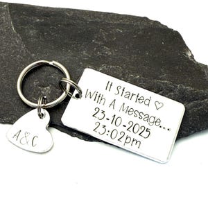 Puede incluir: Un llavero plateado con una etiqueta rectangular y un dije en forma de coraz&oacute;n. La etiqueta rectangular dice "It Started With A Message..." con una fecha y hora. El dije de coraz&oacute;n tiene las iniciales "A&C".