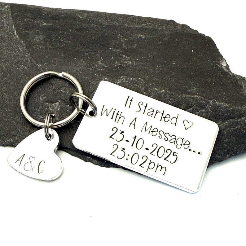Puede incluir: Un llavero plateado con una etiqueta rectangular y un dije en forma de coraz&oacute;n. La etiqueta rectangular dice "It Started With A Message..." con una fecha y hora. El dije de coraz&oacute;n tiene las iniciales "A&C".