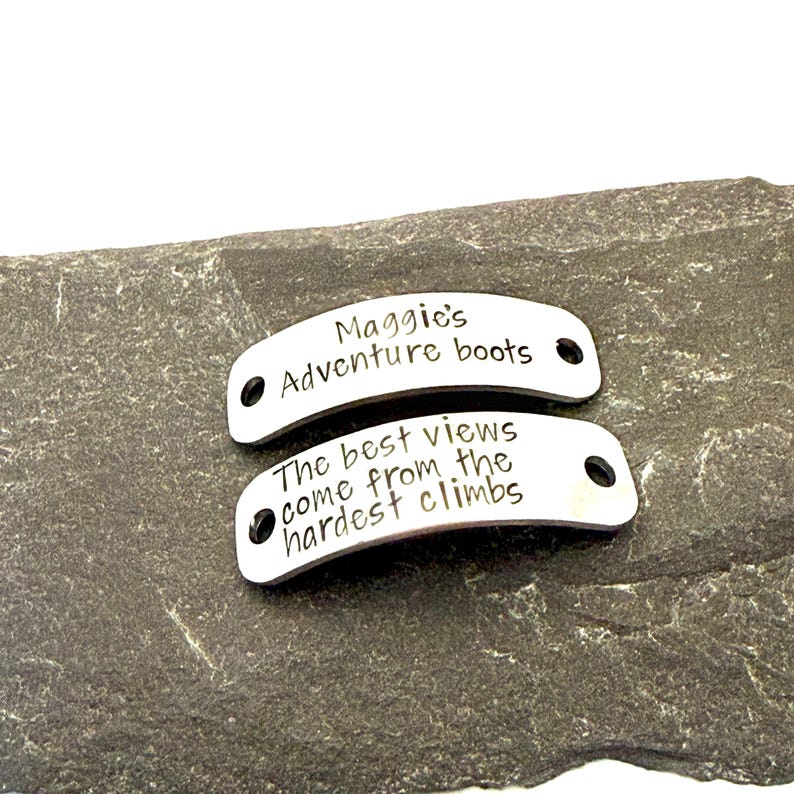 May include: Two silver metal tags with black text. The top tag reads "Maggie's Adventure boots." The bottom tag says "The best views come from the hardest climbs." Both tags have a hole on each end.