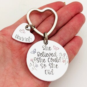 Pode incluir: Um porta-chaves de metal prateado com um anel em forma de cora&ccedil;&atilde;o, um pequeno pendente em forma de cora&ccedil;&atilde;o gravado com o nome Hannah e um &iacute;cone de cora&ccedil;&atilde;o, e um pendente circular maior gravado com o texto She Believed She Could So She Did e flores.