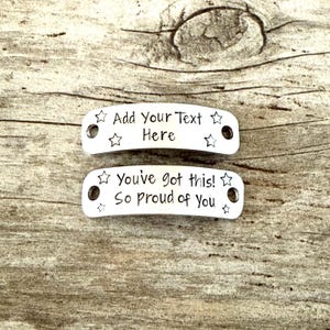 May include: Two silver metal tags with rounded edges and star accents. The top tag reads "Add Your Text Here." The bottom tag says "You've got this! So proud of You." Both tags have holes for attaching.
