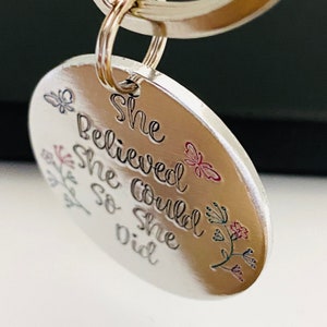 Pode incluir: Um porta-chaves redondo de metal prateado com o texto gravado She Believed She Could So She Did. O pendente est&aacute; decorado com pequenos motivos florais e de borboletas gravados e coloridos em tons de rosa, azul e verde.