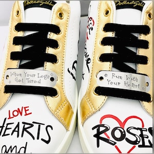 May include: White sneakers with gold accents and black laces. The shoes feature the text "When Your Legs Get Tired" and "Run With Your Heart" on silver tags. The design includes red and black text with hearts and roses.