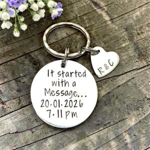 May include: A silver-coloured metal keyring featuring a circular disc and a smaller heart-shaped charm. The circular disc is engraved with the text "It started with a Message... 20.01.2026 7:11 pm" and the heart charm is engraved with the letters "R & C".