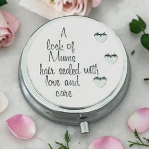 May include: A silver-colored, round keepsake box with the inscription "A lock of Mums hair sealed with love and care." The lid features three heart-shaped cutouts. Pink rose petals and greenery surround the box.