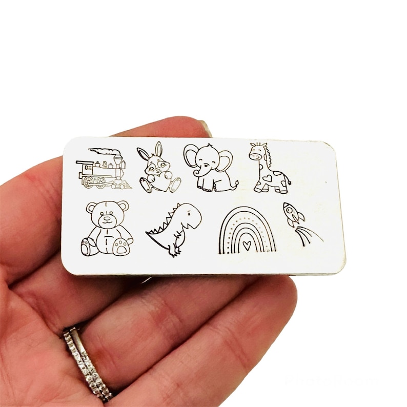 Puede incluir: Un sello met&aacute;lico rectangular con ilustraciones grabadas de un tren, un conejo, un elefante, una jirafa, un oso de peluche, un dinosaurio, un arco&iacute;ris y un cohete. Las im&aacute;genes son en blanco y negro.