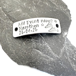 May include: A silver metal tag with the text "Add Event name? Marathon 26.04.26" and two pairs of sneakers. The tag has two holes and star designs. The tag is on a gray stone surface.