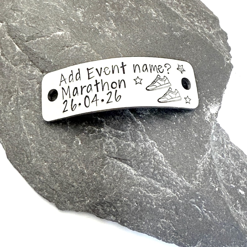 May include: A silver metal tag with the text "Add Event name? Marathon 26.04.26" and two pairs of sneakers. The tag has two holes and star designs. The tag is on a gray stone surface.