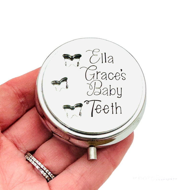 Puede incluir: Caja redonda plateada para guardar dientes de beb&eacute;. La tapa presenta tres recortes con forma de diente y la inscripci&oacute;n "Ella Grace's Baby Teeth" en escritura elegante. Se ve una peque&ntilde;a bisagra y cierre plateados.
