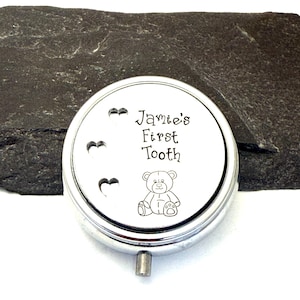 Puede incluir: Una caja redonda de recuerdo de color plateado con el texto "Jamie's First Tooth" y una ilustraci&oacute;n de un osito de peluche. Tres recortes en forma de coraz&oacute;n est&aacute;n en el lado izquierdo. La caja tiene un peque&ntilde;o asa plateada.
