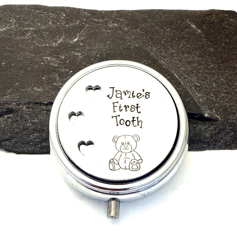 Puede incluir: Una caja redonda de recuerdo de color plateado con el texto "Jamie's First Tooth" y una ilustraci&oacute;n de un osito de peluche. Tres recortes en forma de coraz&oacute;n est&aacute;n en el lado izquierdo. La caja tiene un peque&ntilde;o asa plateada.
