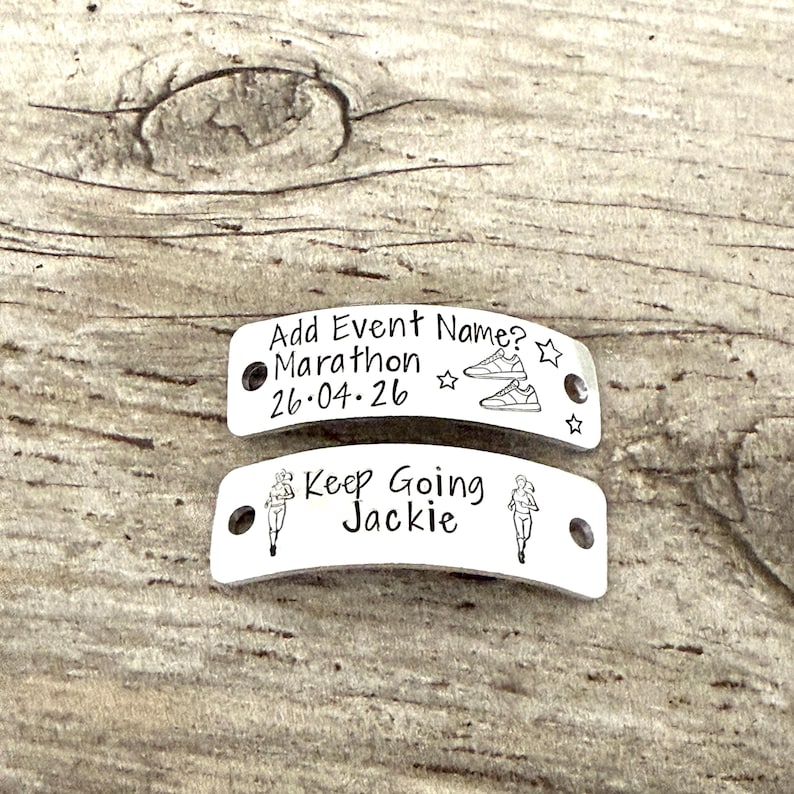 May include: Two silver-colored metal tags with engraved text. The top tag reads "Add Event Name? Marathon 26.04.26" with shoe and star illustrations. The bottom tag says "Keep Going Jackie" with runner illustrations.