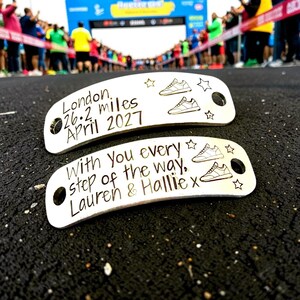May include: Two silver-toned metal shoe tags with rounded ends and holes for laces. The top tag reads London, 26.2 miles, April 2027 with two sneaker icons and stars. The bottom tag reads With you every step of the way, Lauren & Hallie x with sneakers.