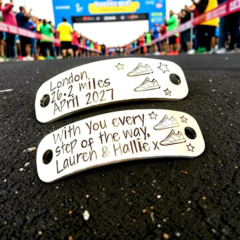 May include: Two silver-toned metal shoe tags with rounded ends and holes for laces. The top tag reads London, 26.2 miles, April 2027 with two sneaker icons and stars. The bottom tag reads With you every step of the way, Lauren & Hallie x with sneakers.