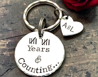 以下が含まれることがあります： 「Years & Counting...」と書かれた丸いタグと「A&L」のイニシャルが入ったハート型のタグが付いた、シルバーカラーのキーホルダー。キーホルダーには、鍵用の大きなリングも付いています。背景には赤いバラがあります。