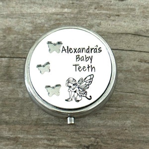 Puede incluir: Una caja redonda plateada para guardar dientes de beb&eacute;. La tapa presenta el texto "Alexandra's Baby Teeth" con tres recortes de mariposas y una ilustraci&oacute;n de hada. La caja est&aacute; sobre una superficie de madera.